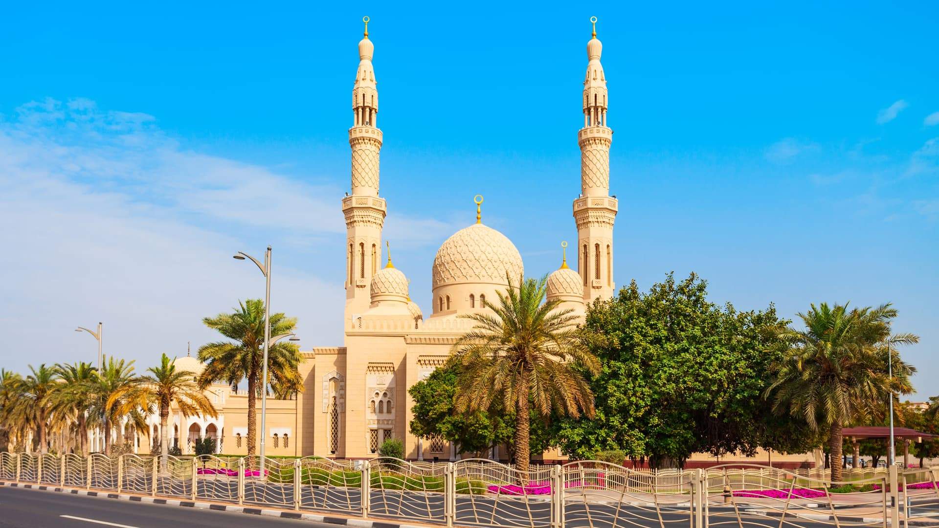 Daytime view of the iconic Jumeirah Mosque in Dubai, showcasing its elegant cream-coloured domes and twin minarets. Surrounded by palm trees and lush greenery, the mosque stands against a clear blue sky, reflecting traditional Islamic architecture.