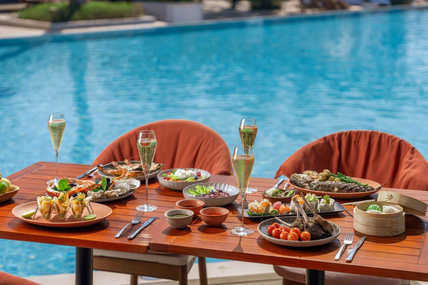 A luxurious poolside dining setup at Banyan Tree Dubai featuring an elegant wooden table adorned with gourmet dishes including seafood, grilled lamb chops, sushi, salads, and champagne glasses, with a sparkling blue pool in the background.