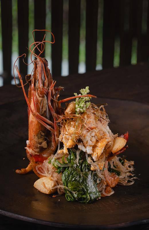 A beautifully plated Thai seafood dish featuring a grilled river prawn with its shell intact, served atop glass noodles, fresh greens, crispy garlic, and aromatic herbs, presented on a dark ceramic plate with a rustic wooden backdrop.