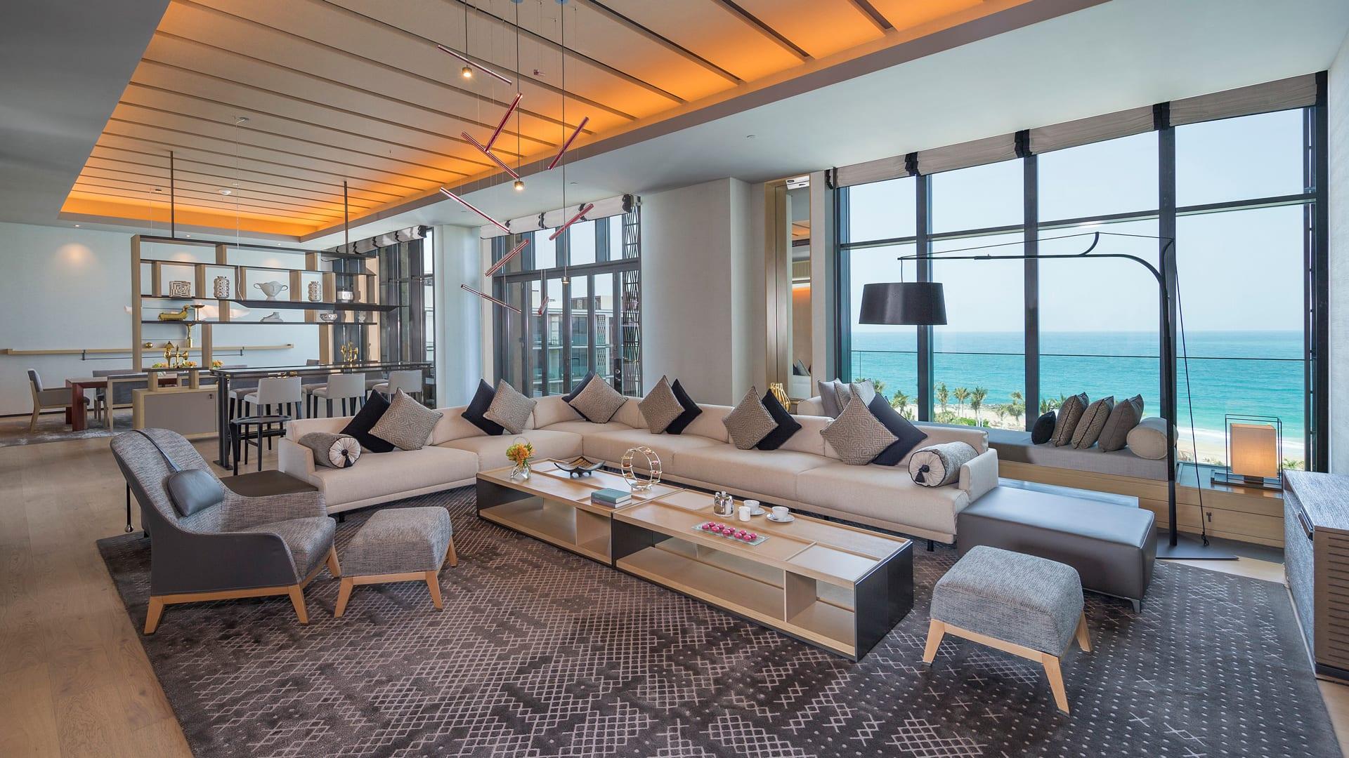 Luxurious modern living room in an oceanfront royal suite featuring a large beige sectional sofa with black and grey cushions, a stylish coffee table with refreshments, contemporary lighting fixtures, floor-to-ceiling windows offering a panoramic sea view, and elegant decor throughout.