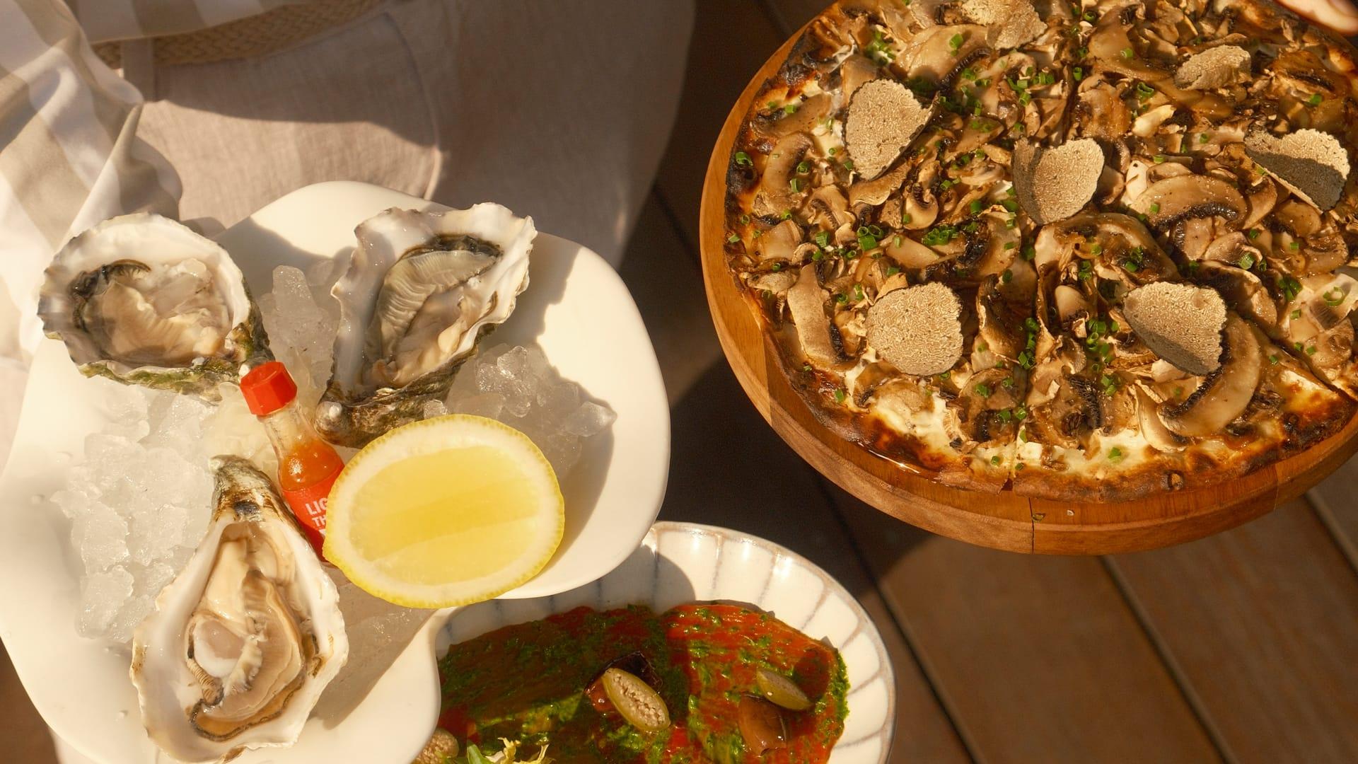 A top-down view of a gourmet meal featuring fresh oysters on ice with lemon and hot sauce, a truffle mushroom pizza on a wooden board, and a vibrant dish with green herbs and red sauce, all set on a wooden table in natural sunlight.