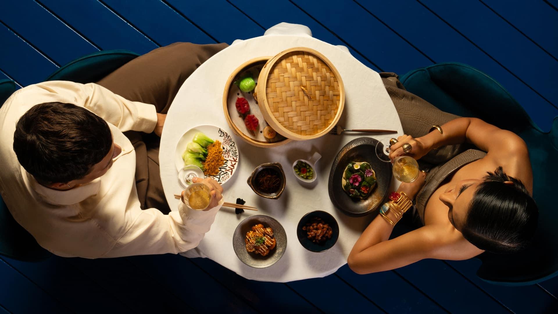 A stylish couple is dining at a round table set with an elegant Asian-inspired meal, including dim sum in a bamboo steamer, vibrant vegetables, and artistic plating. They are seated on green velvet chairs, toasting with champagne, captured from an overhead view on a blue wooden floor.