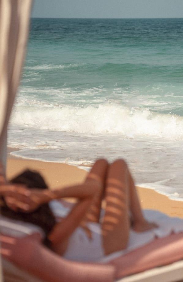 Relaxed beachside scene with a woman lounging on a sunbed under soft cabana shadows, facing gentle waves washing onto the sandy shore. The ocean view and warm tones evoke a tranquil, carefree atmosphere.