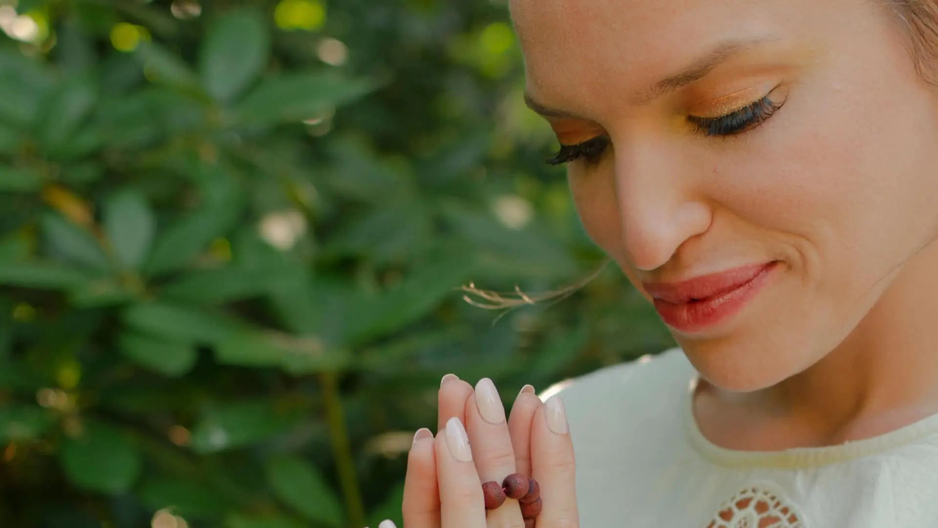 Close-up of a peaceful woman with eyes closed, smiling gently while holding prayer beads in a meditative gesture. She stands in front of lush green foliage, radiating calm and mindfulness in a natural outdoor setting.