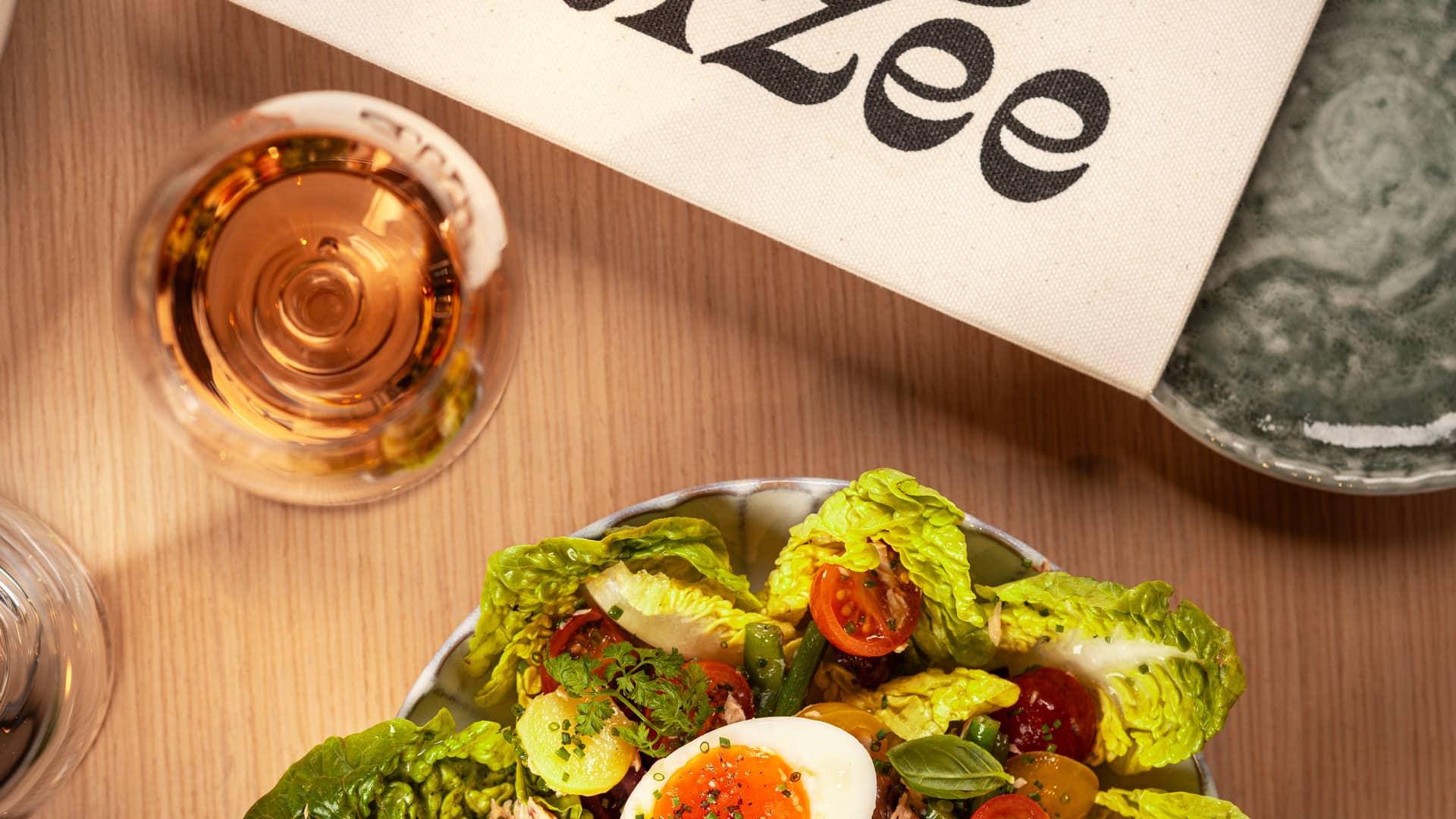 Top-down view of a vibrant salad with cherry tomatoes, crisp lettuce, green beans, herbs, and a soft-boiled egg, paired with a glass of rosé wine. A fabric napkin with 'Alizée' branding is partially visible on a wooden table, creating a fresh and inviting dining scene.
