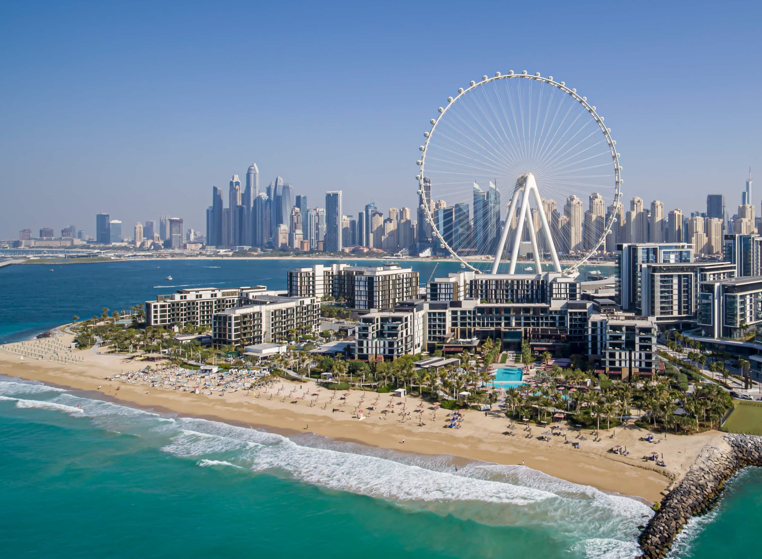 Aerial view of a luxury beachfront resort on Bluewaters Island, Dubai, with Ain Dubai observation wheel towering in the background. The turquoise sea meets a sandy beach lined with sunbeds, while the modern skyline of Dubai Marina creates a dramatic urban contrast.