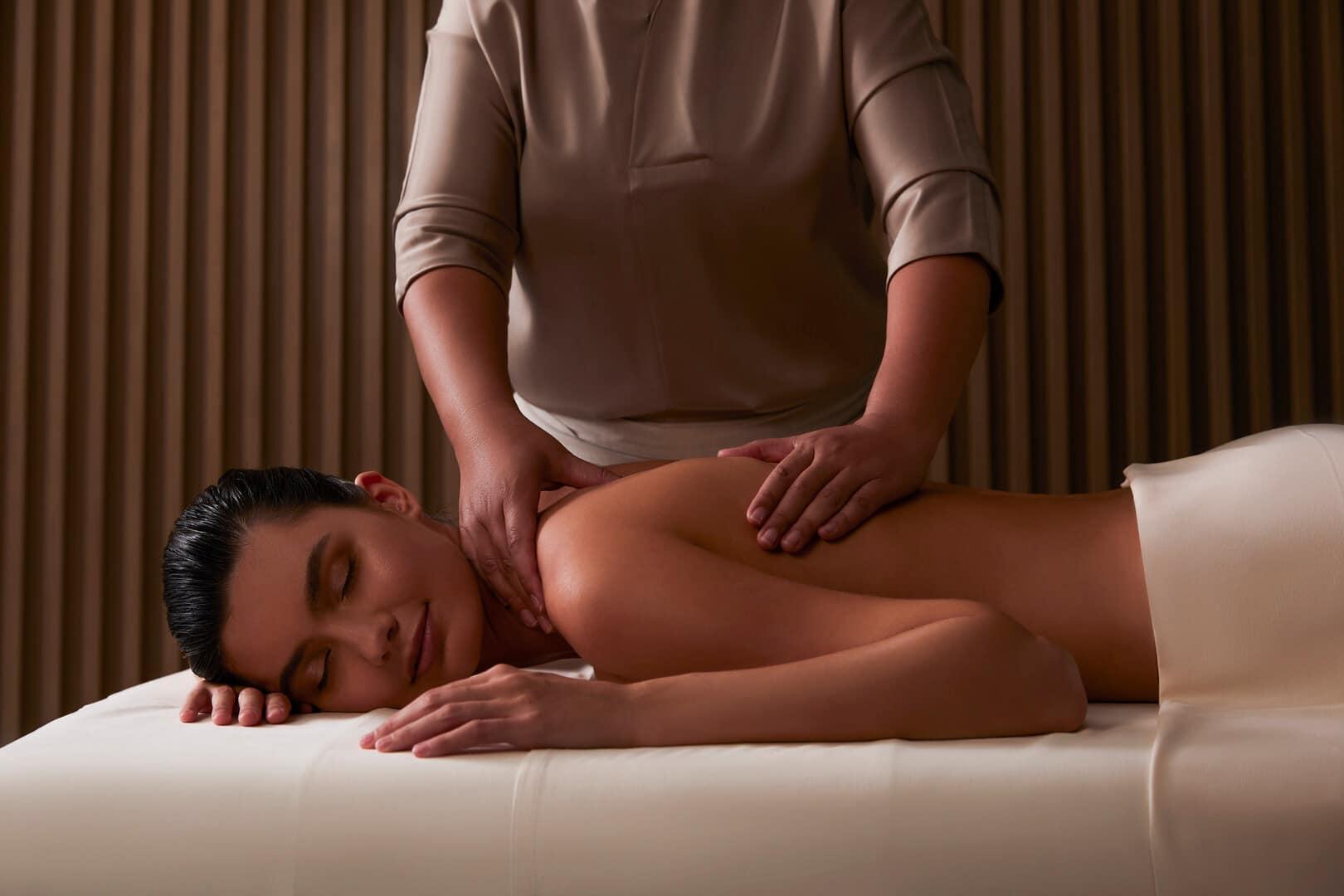 Serene spa setting with a woman lying on a massage table receiving a relaxing back and shoulder massage from a therapist. The tranquil ambiance is enhanced by soft lighting and a warm, neutral-toned interior, creating a calming wellness experience.