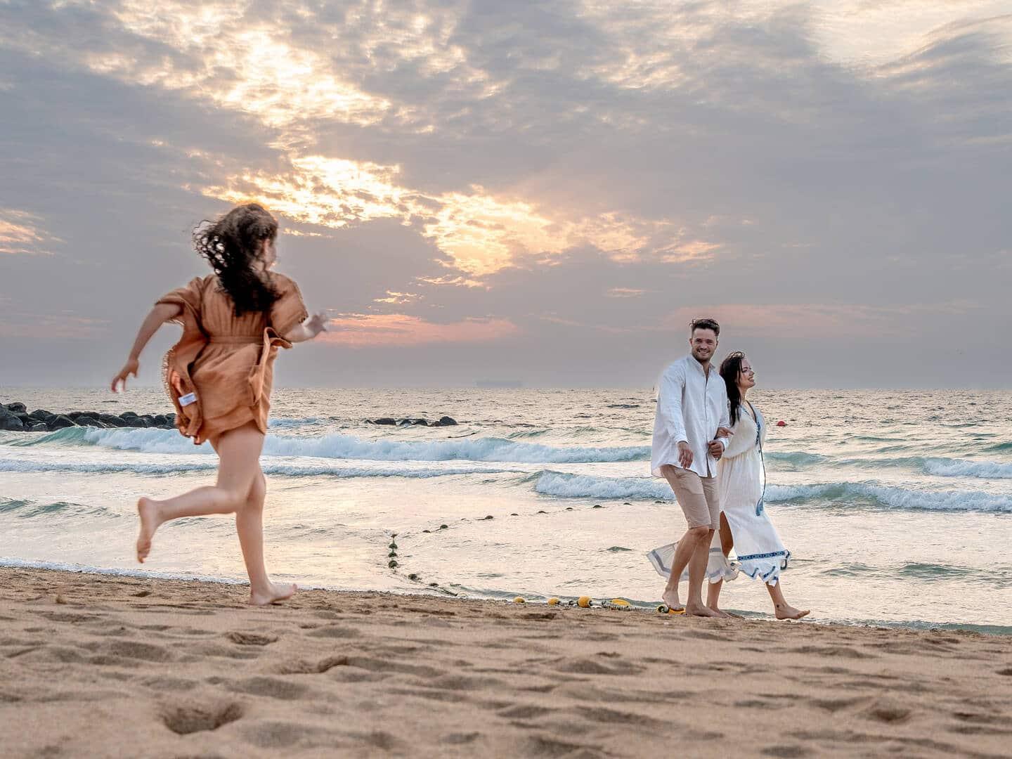 Joyful beach scene at sunset with a couple walking hand-in-hand along the shoreline, smiling and relaxed. A young girl runs toward them barefoot across the sand, capturing a moment of family connection against the backdrop of gentle waves and a dramatic, colourful sky.