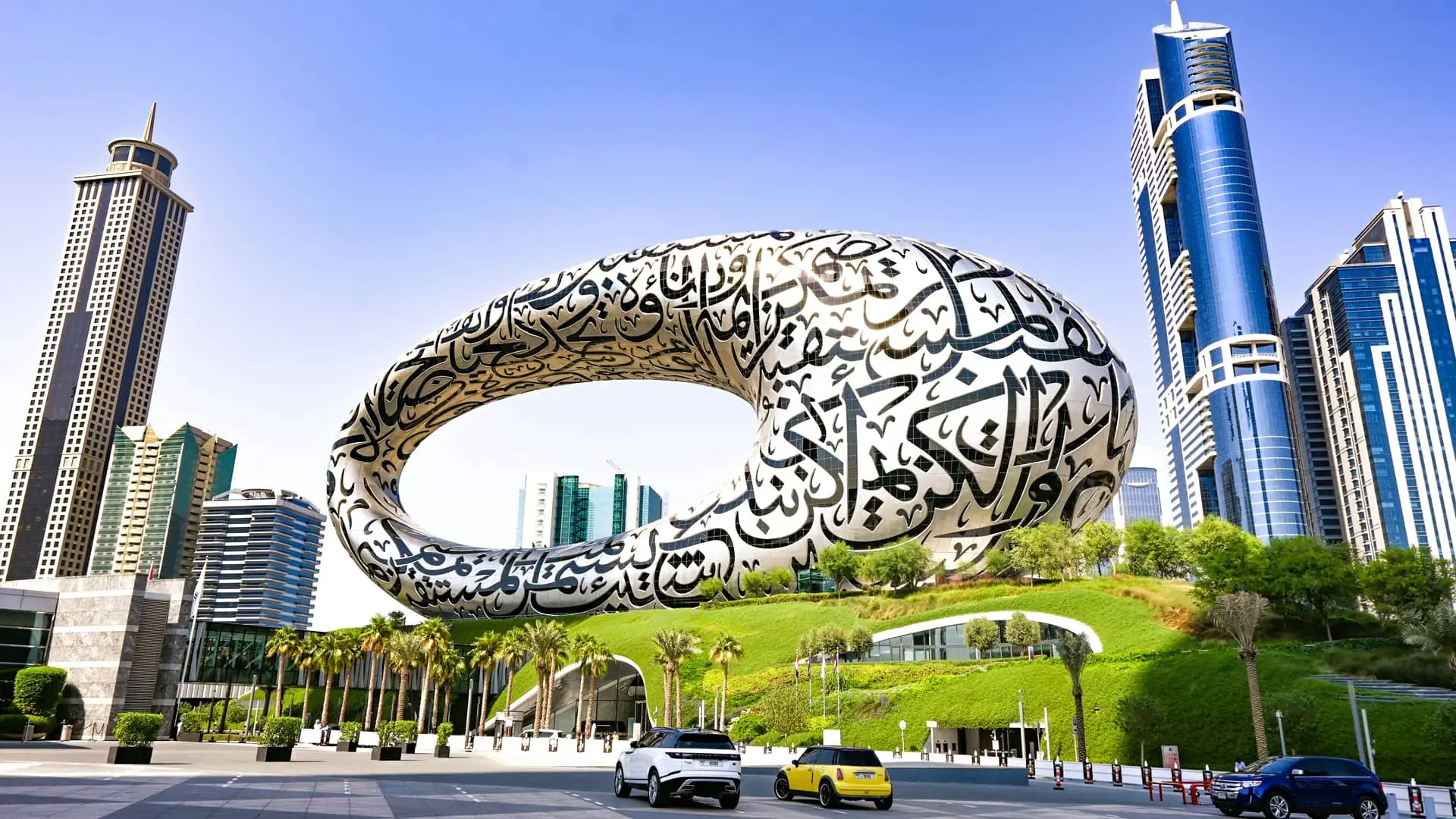 Daytime view of the Museum of the Future in Dubai, a striking torus-shaped structure adorned with flowing Arabic calligraphy. Surrounded by modern skyscrapers and palm trees, the futuristic building sits atop a green landscaped mound under a clear blue sky.