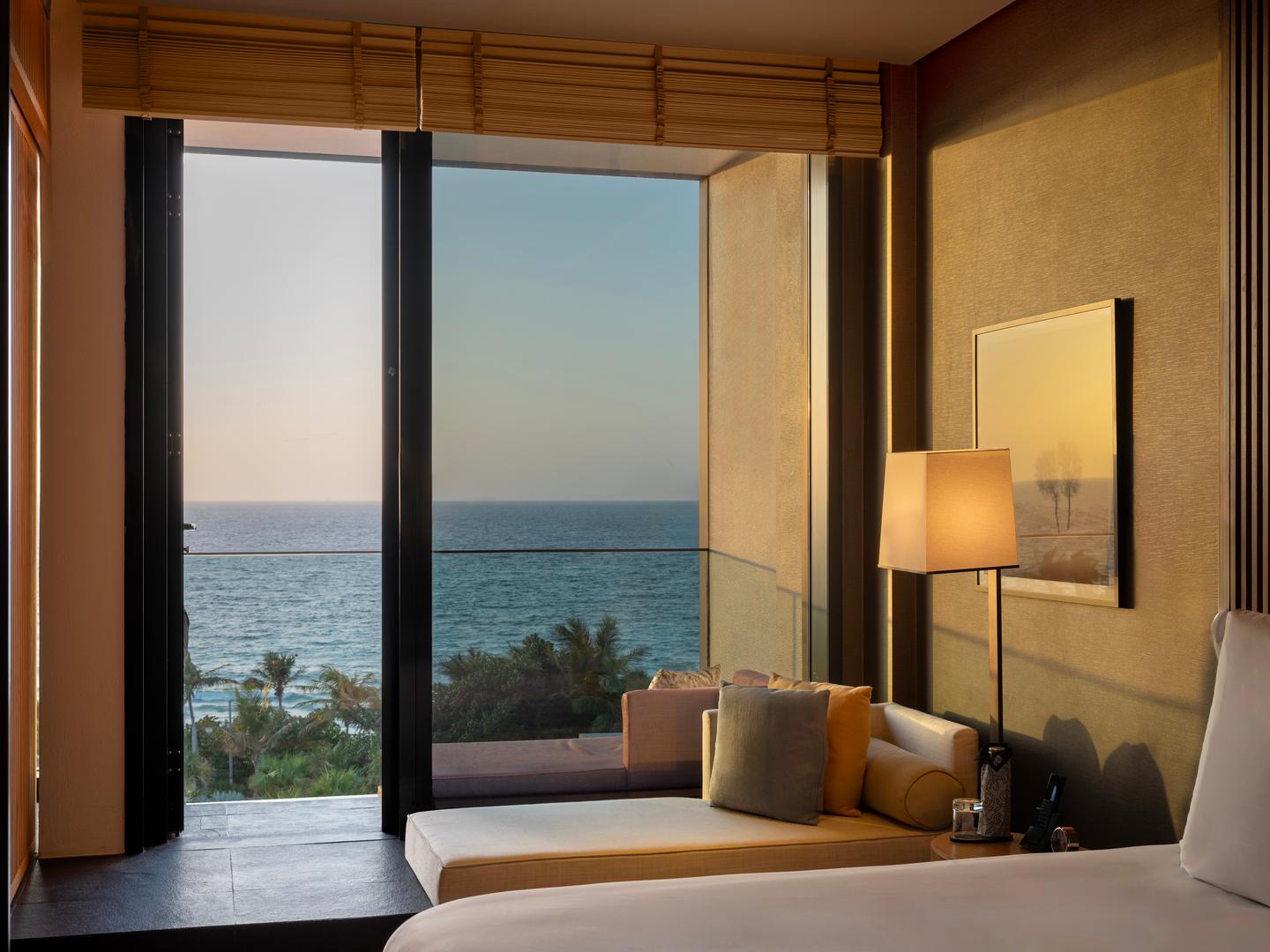 Luxurious oceanview guestroom with a contemporary lounge corner, floor-to-ceiling windows, and panoramic views of the sea and palm trees at sunset.