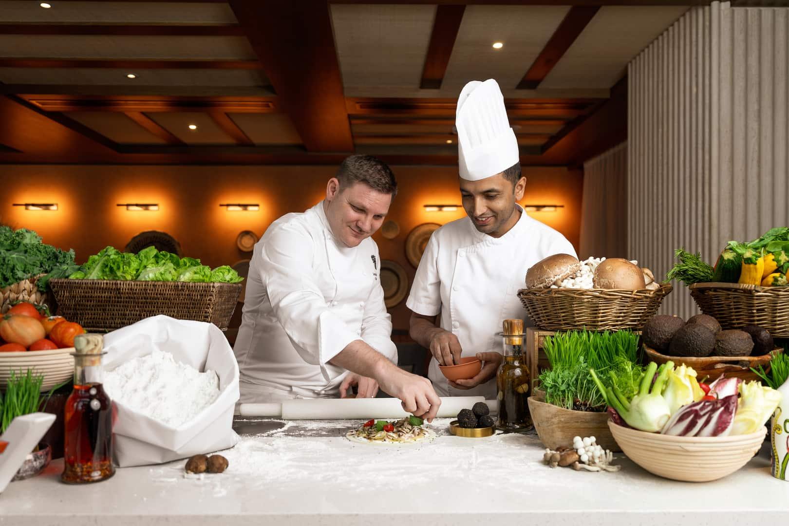 Two chefs at Banyan Tree Dubai Culinary Academy preparing a gourmet dish together in a warmly lit kitchen, surrounded by fresh vegetables, herbs, flour, mushrooms, and olive oil on a countertop.