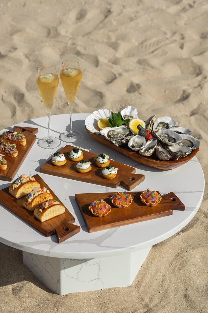 A beachfront table set with gourmet bites including oysters, canapés, and mini tacos, complemented by two glasses of champagne, offering a luxurious seaside dining experience.