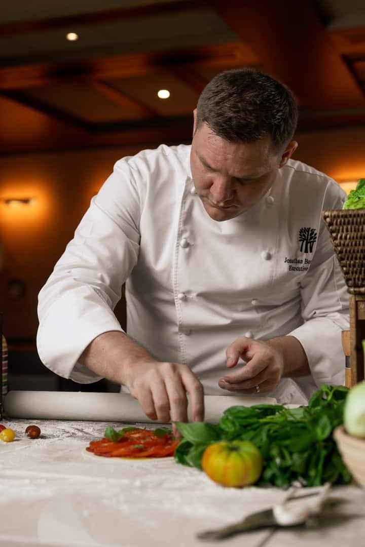 Executive Chef Jonathan Boushall of Banyan Tree Dubai carefully garnishing a tomato-based dish in a warm, ambient kitchen setting, surrounded by fresh herbs and vegetables.