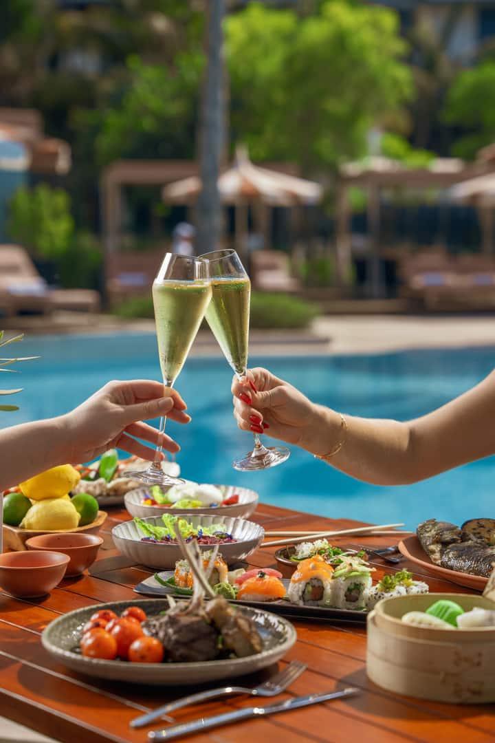 Two people toast with champagne flutes over an elegant poolside lunch featuring sushi, salads, and grilled dishes at Banyan Tree Dubai.