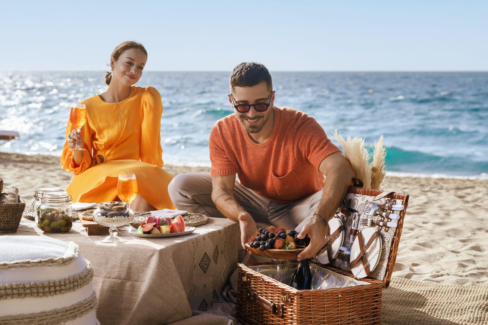 A stylish couple enjoys a luxury beachside picnic with fresh fruits, snacks, and Aperol spritz, seated on a low table with coastal views and a wicker basket setup.
