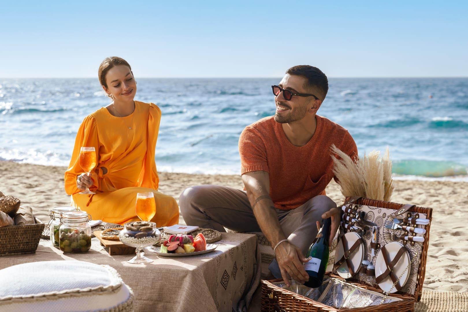 A stylish couple enjoys a luxury beachside picnic with fresh fruits, snacks, and Aperol spritz, seated on a low table with coastal views and a wicker basket setup.