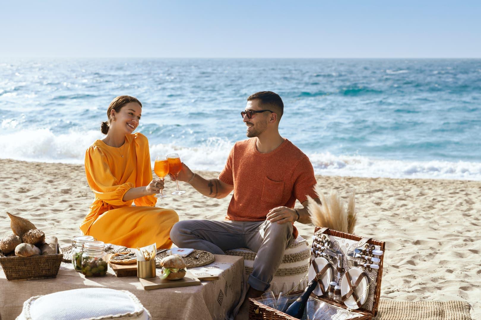A stylish couple enjoys a luxury beachside picnic with fresh fruits, snacks, and Aperol spritz, seated on a low table with coastal views and a wicker basket setup.