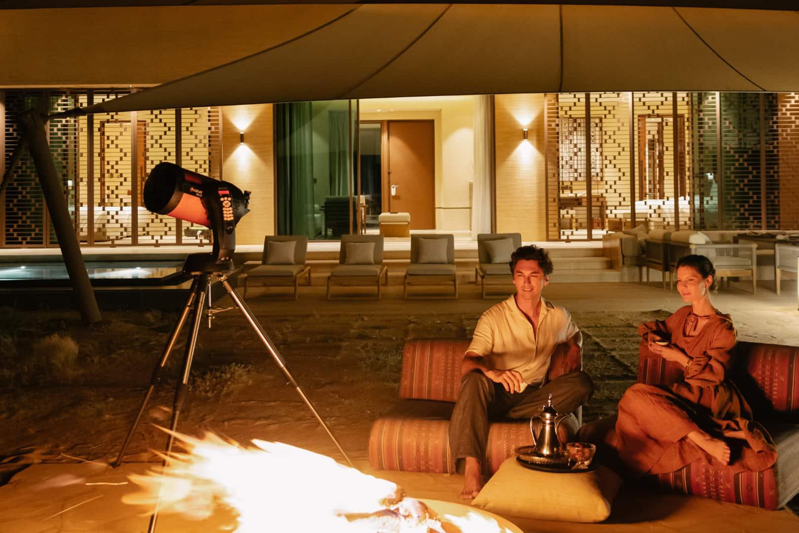 A couple sit by a telescope in front of a fire pit at Banyan Tree AlUla