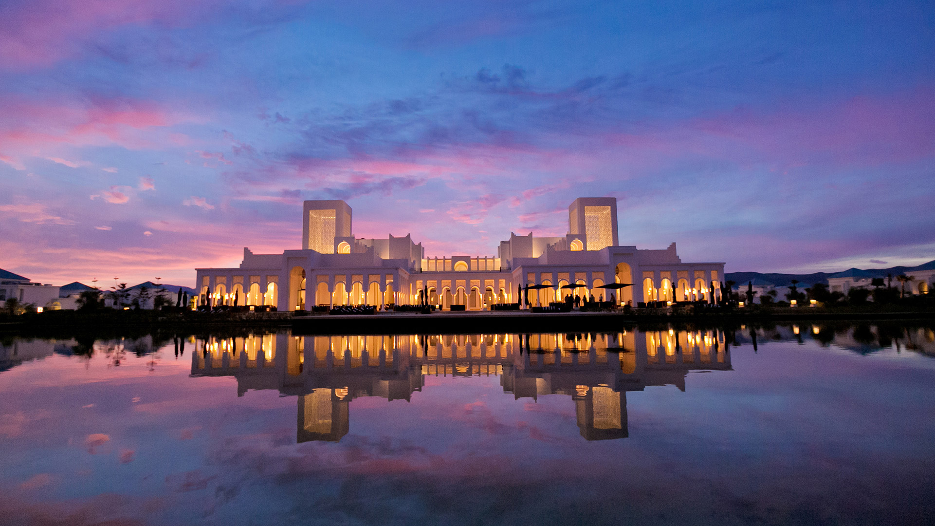 Banyan Tree Tamouda Bay's illuminated facade reflects in tranquil water at twilight.