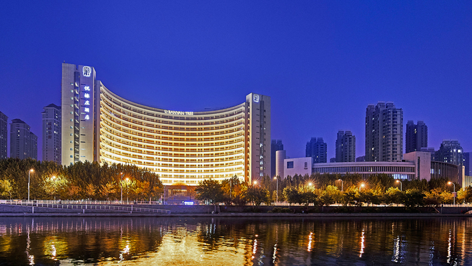 Curved, illuminated building at night; reflection on water; Banyan Tree Tianjin Riverside.