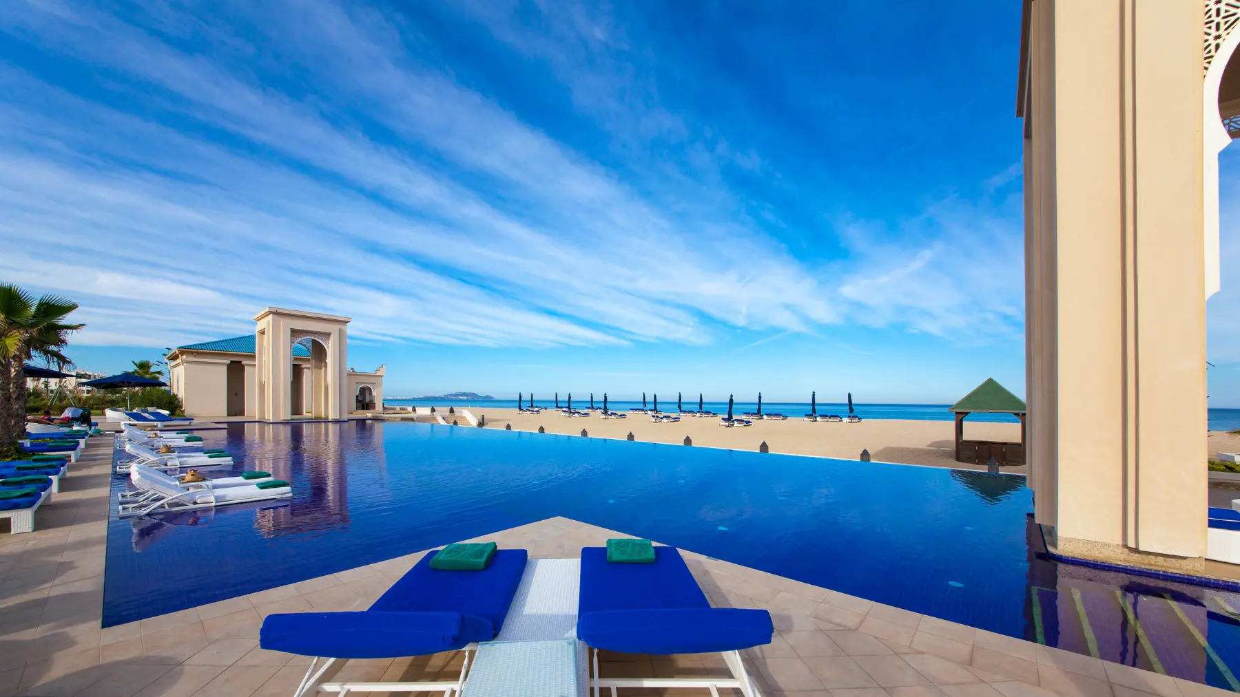 Poolside loungers rest by a tranquil infinity pool overlooking the beach at Banyan Tree Tamouda Bay.