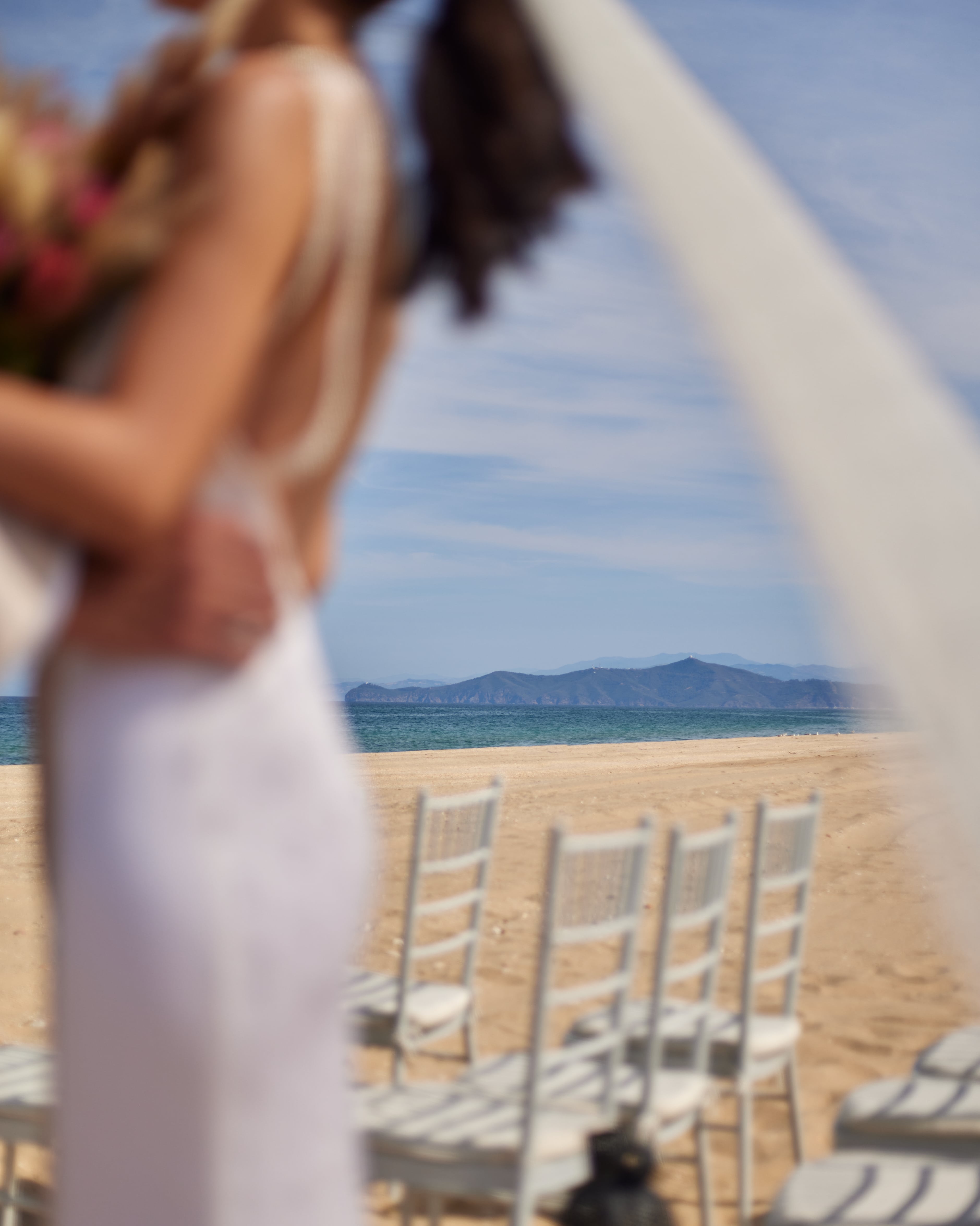 Bride stands on sandy beach with chairs; ocean and mountains in background at Banyan Tree Tamouda Bay wedding.
