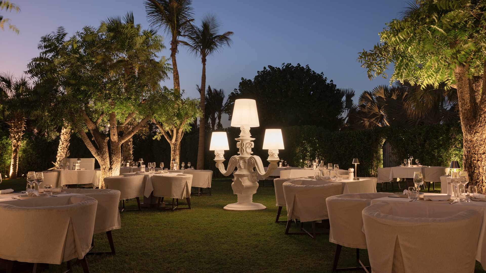 Outdoor dining tables at Blue Door Dubai with elegant lamps adorn lush garden.