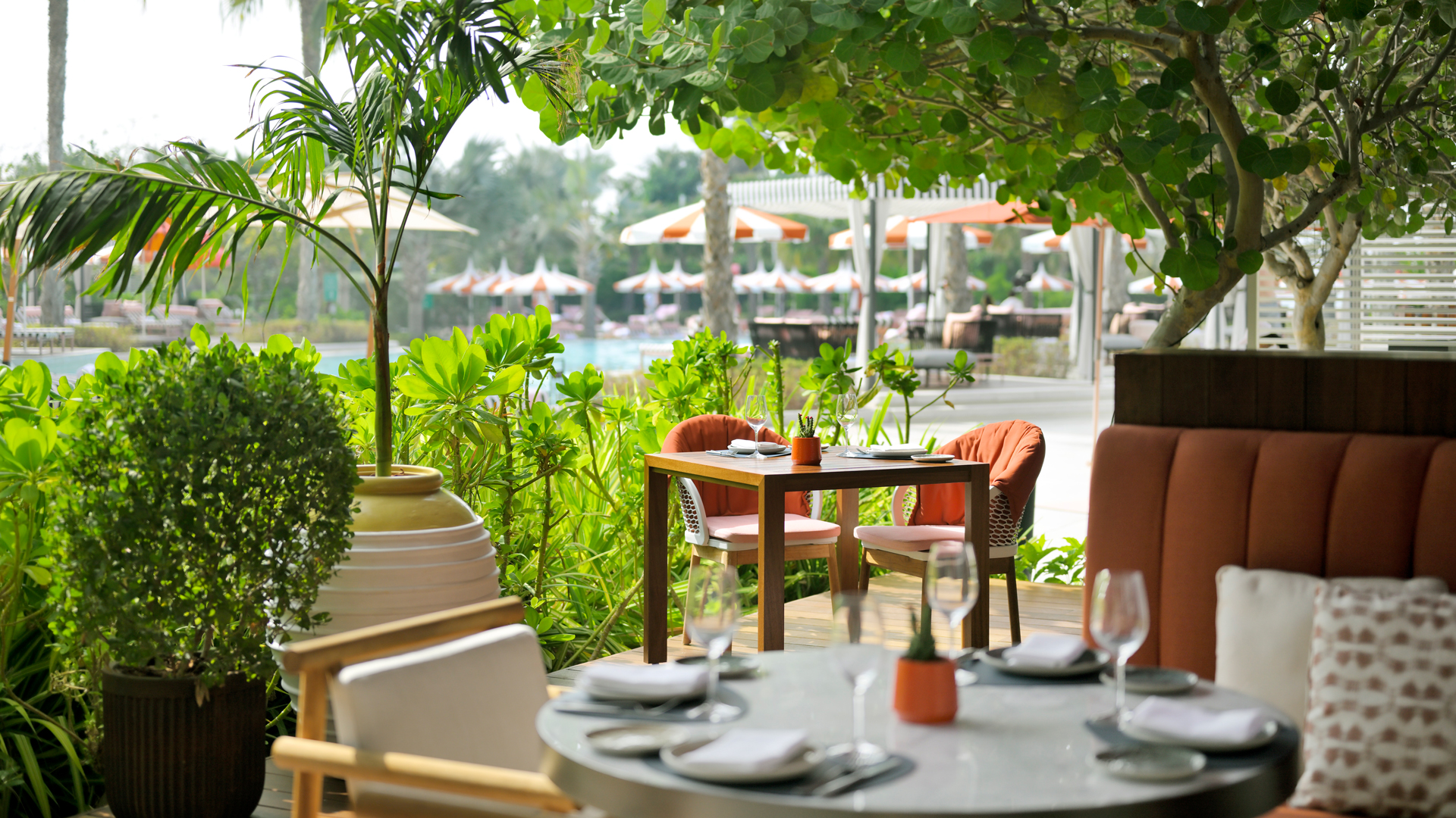 A beautifully set outdoor dining area surrounded by lush greenery, offering a serene ambiance with elegant tables, plush seating, and views of a resort pool with orange-and-white umbrellas in the background.