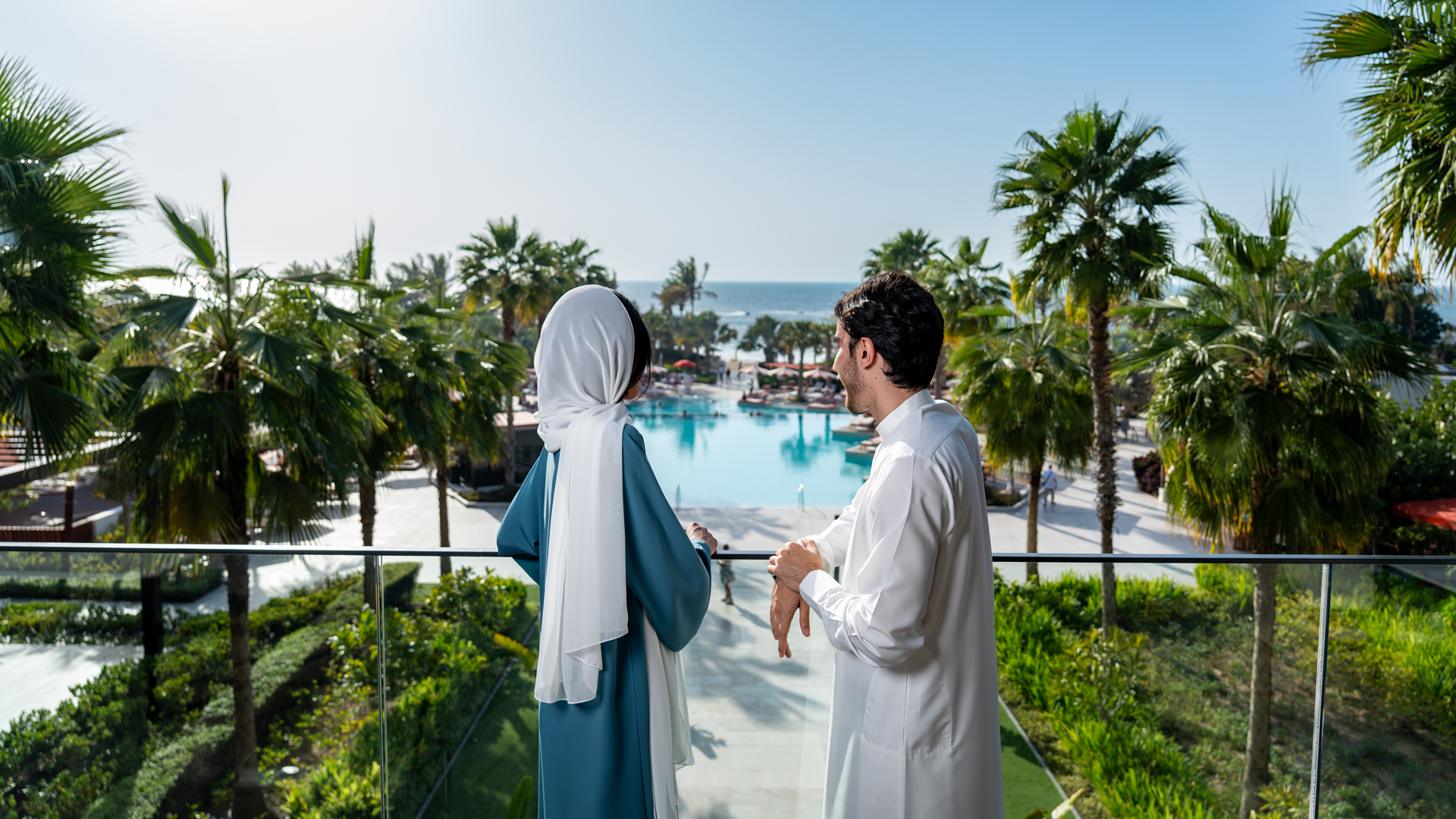 A couple stands on a balcony overlooking a luxurious resort pool surrounded by lush palm trees, with the ocean stretching into the horizon. The woman wears a white headscarf and a flowing teal dress, while the man wears a traditional white thobe, both enjoying the serene and picturesque view.