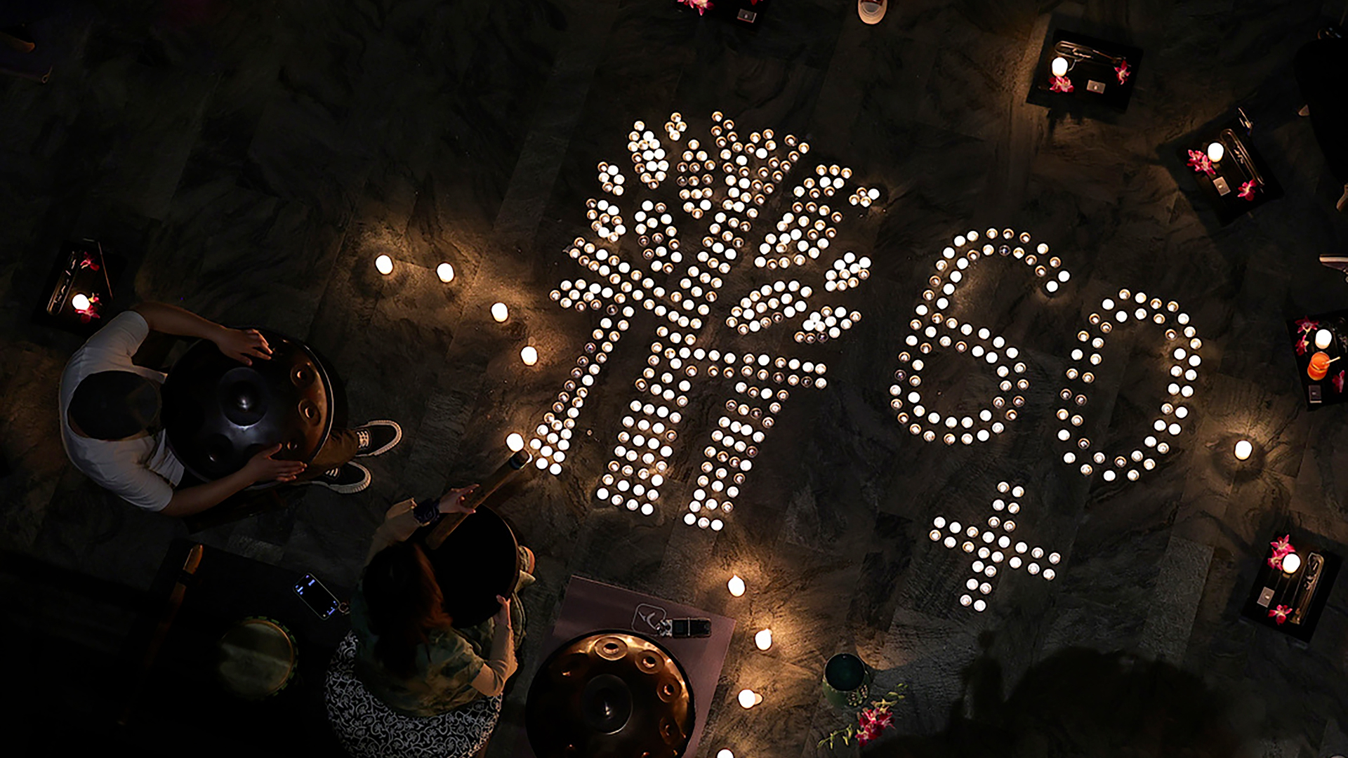 Earth Hour 2025 A nighttime aerial view of a candle-lit display featuring a tree symbol and the number "60+" arranged on a dark surface. The setting appears to be an Earth Hour event, with individuals playing handpans and other instruments nearby. The warm glow of the candles contrasts with the dimly lit surroundings, creating a serene and symbolic atmosphere.