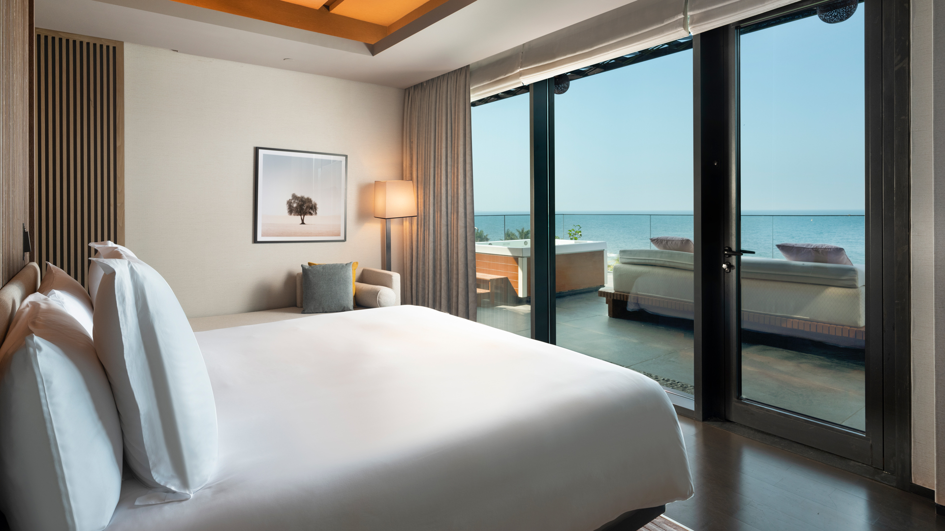 A luxurious oceanfront bedroom featuring a plush king-size bed with crisp white linens, modern décor, and a private terrace with lounge seating and a jacuzzi, offering breathtaking views of the sea through floor-to-ceiling glass doors.