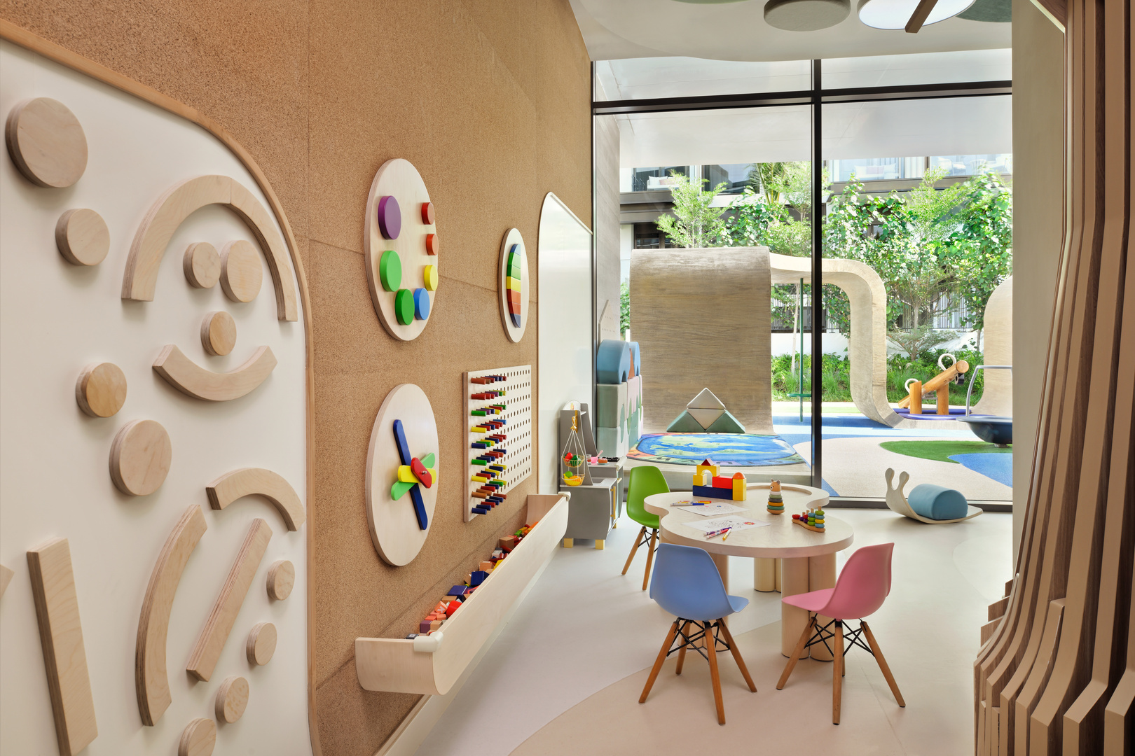 A modern and inviting children's playroom with interactive wooden wall games, colorful chairs, and activity tables. The space features educational toys, a clock learning board, and a pegboard with colorful beads. Large windows offer a view of an outdoor play area with greenery, curved play structures, and a soft play mat.