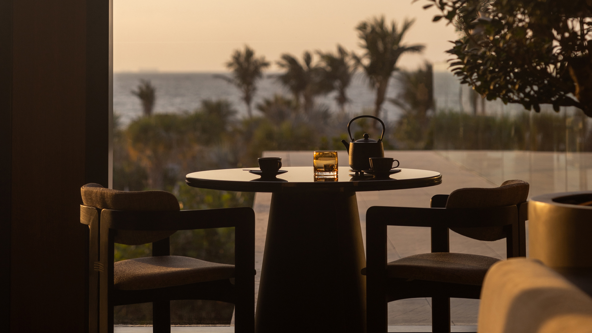Elegant indoor seating setup featuring a round table with two chairs, a teapot, cups, and a candle, set against a serene backdrop of palm trees and ocean views at sunset, creating a warm and tranquil ambiance.
