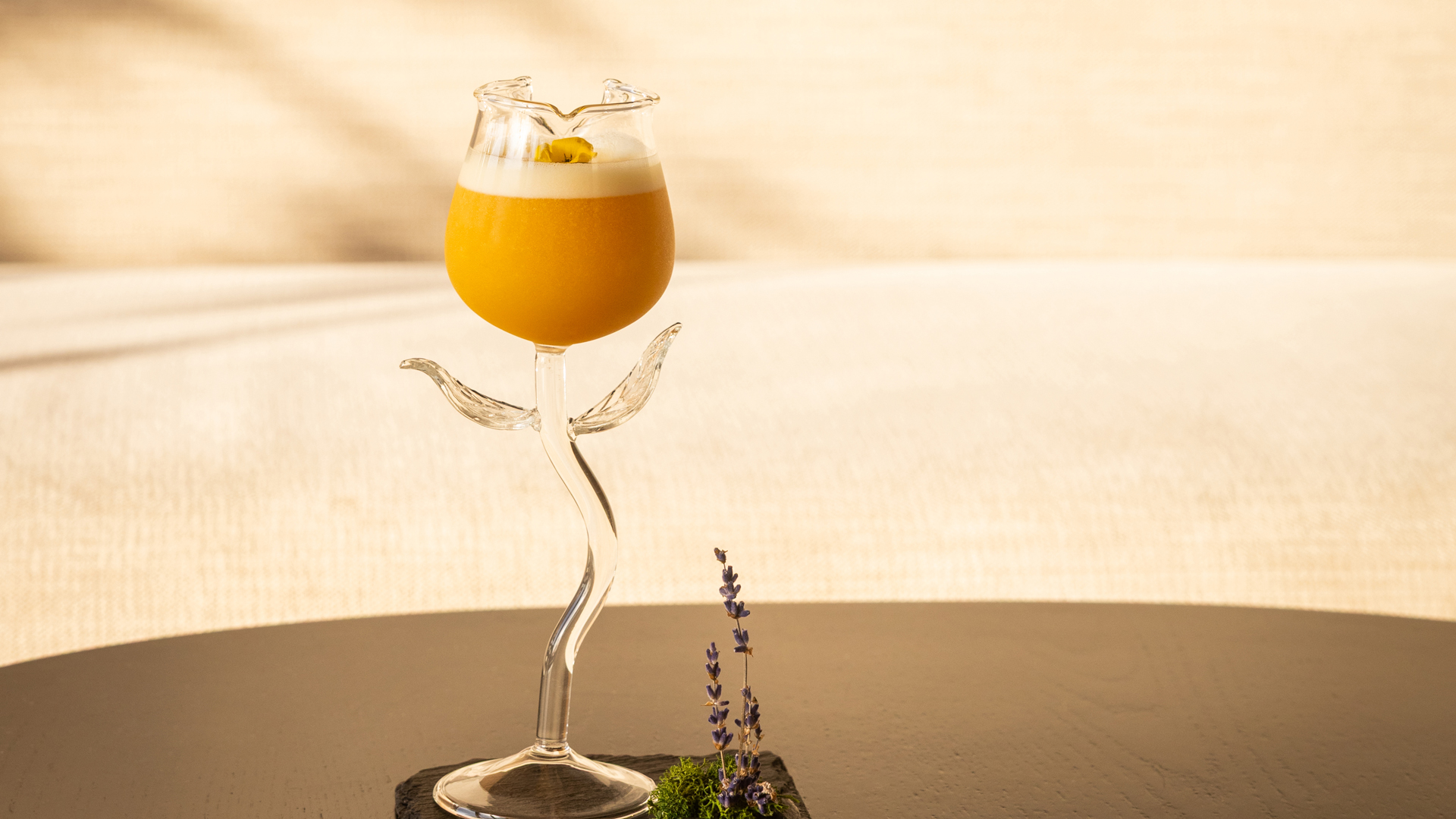 Elegant cocktail served in a sculpted glass shaped like a blooming flower, featuring a vibrant orange drink with a foamy top and edible flower garnish. The glass sits on a dark surface with a small lavender sprig and moss decoration, set against a soft, neutral background.
