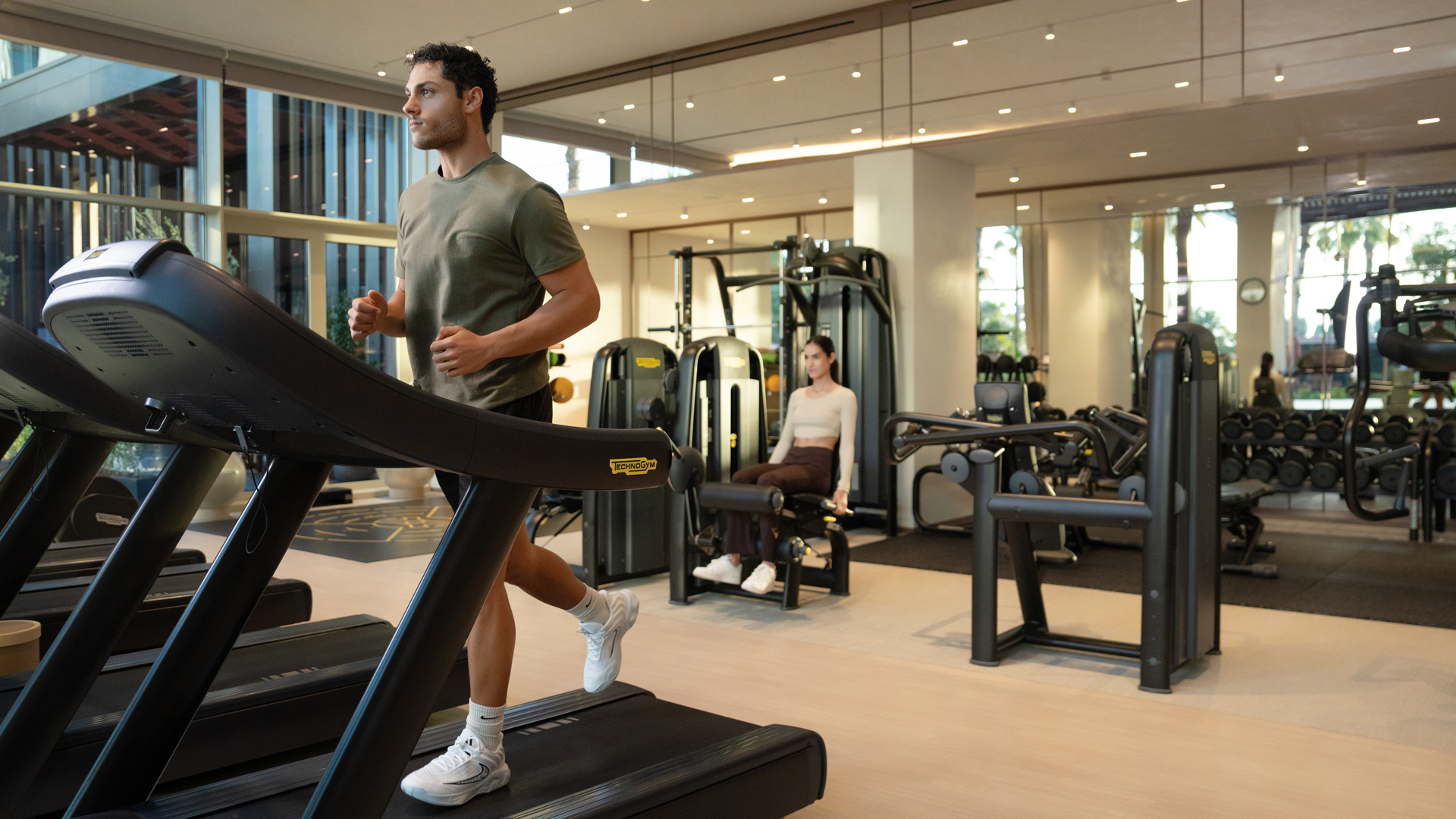 Modern fitness centre with natural lighting and large windows. A man is jogging on a treadmill while a woman uses a leg extension machine in the background. The gym is equipped with Technogym machines and free weights, creating a clean and energising workout environment.
