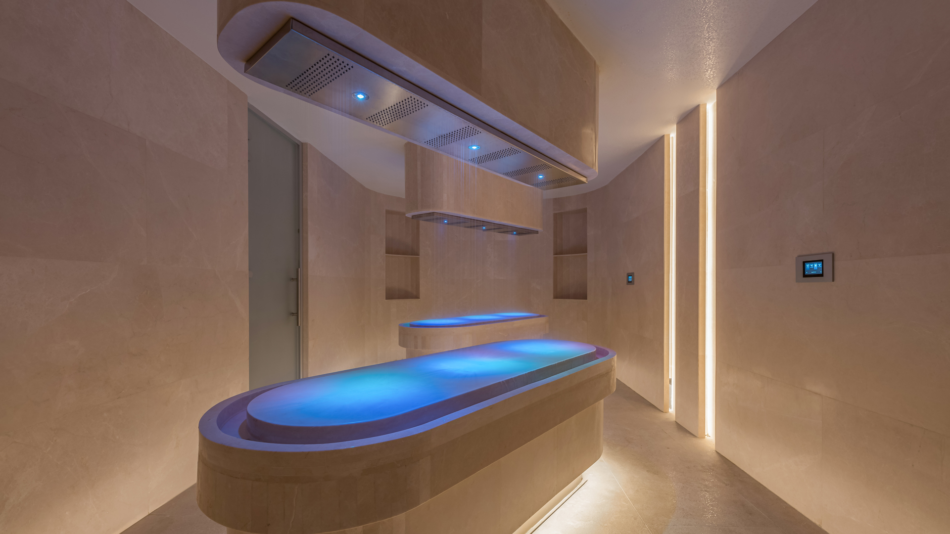 A luxurious spa treatment room featuring a modern rainmist therapy bed illuminated with soft blue lighting. Overhead rain shower panels are suspended above the bed, surrounded by sleek beige stone walls and vertical ambient lighting for a calming atmosphere.