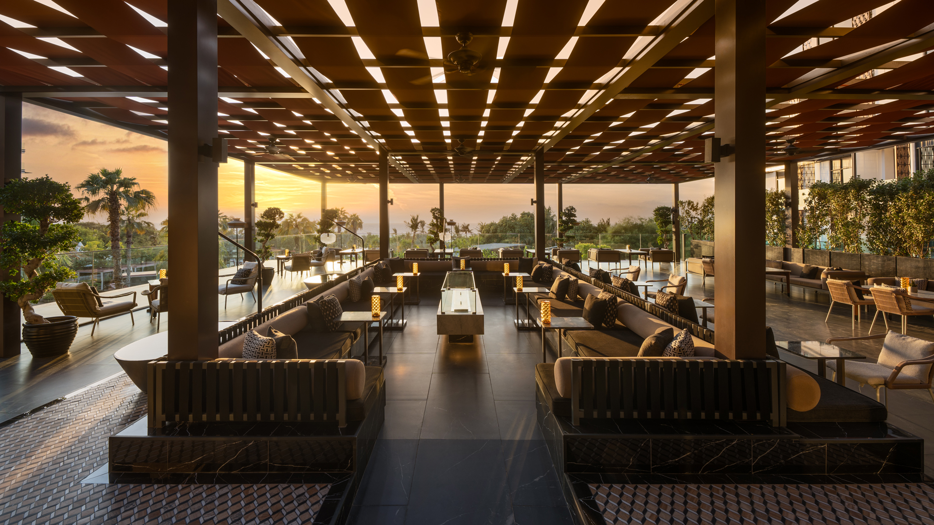 Tocha Stylish rooftop lounge with modern seating, ambient lighting, and a sleek geometric ceiling design. The space features low sofas, elegant tables with glowing lanterns, and panoramic views of palm trees and the ocean at sunset, creating a warm and sophisticated atmosphere.
