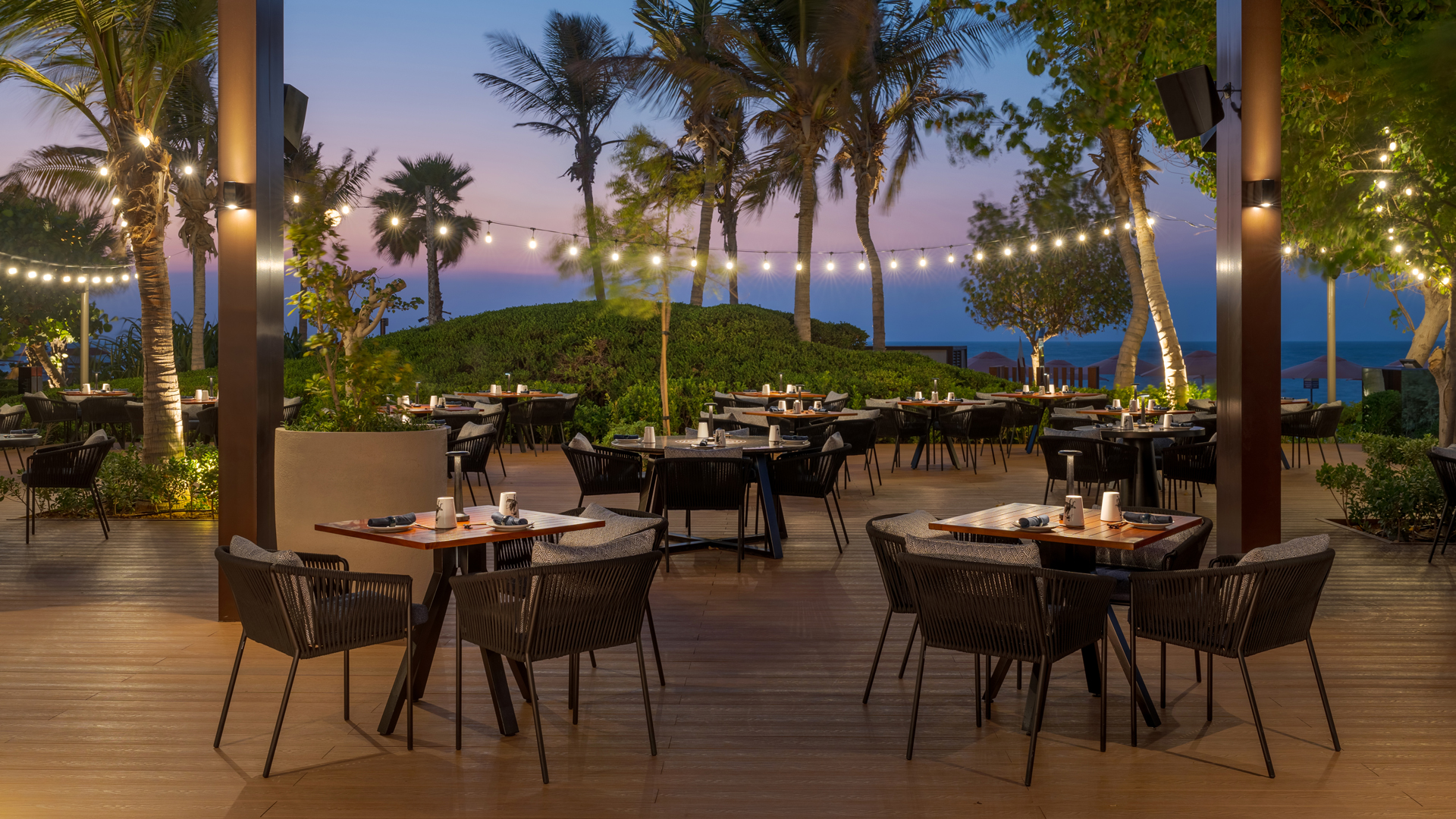 Outdoor beachfront restaurant terrace at sunset, featuring wooden flooring, modern black woven chairs, and neatly set dining tables. String lights hang between palm trees, creating a warm, inviting ambiance with the sea and evening sky in the background.