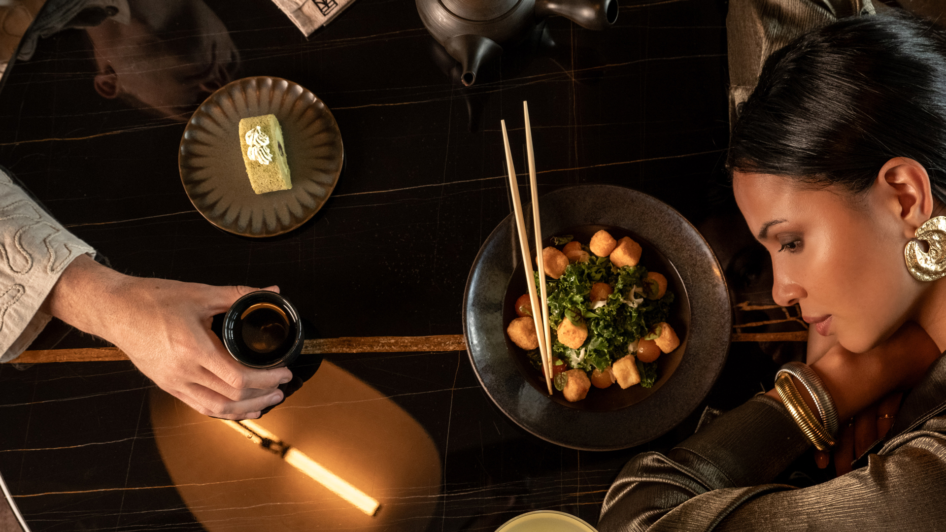 Stylish dining scene captured from above, featuring a woman leaning on a glossy black table set with a salad bowl, chopsticks, a teacup, a teapot, and a dessert plate with matcha cake. The setup highlights a sophisticated, intimate moment with elegant accessories and warm ambient lighting.