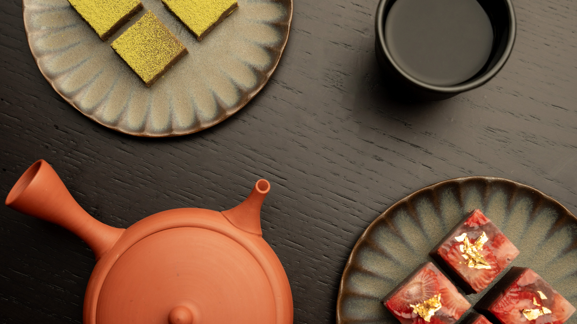 Flat lay of an elegant tea setup featuring a traditional clay teapot, a cup of tea, and two ceramic plates — one with green matcha-dusted sweets and the other with strawberry chocolate squares topped with gold leaf, all placed on a dark wooden surface.
