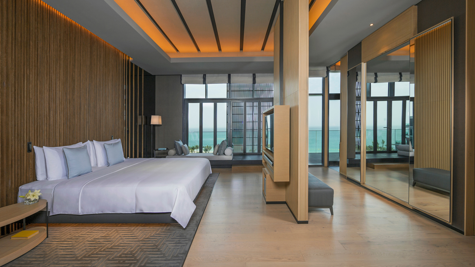 Elegant oceanfront bedroom in a luxury suite featuring a large bed with white linens and soft blue accent pillows, wooden paneling, modern lighting, a built-in TV divider, mirrored wardrobes, and floor-to-ceiling windows offering a stunning view of the sea.