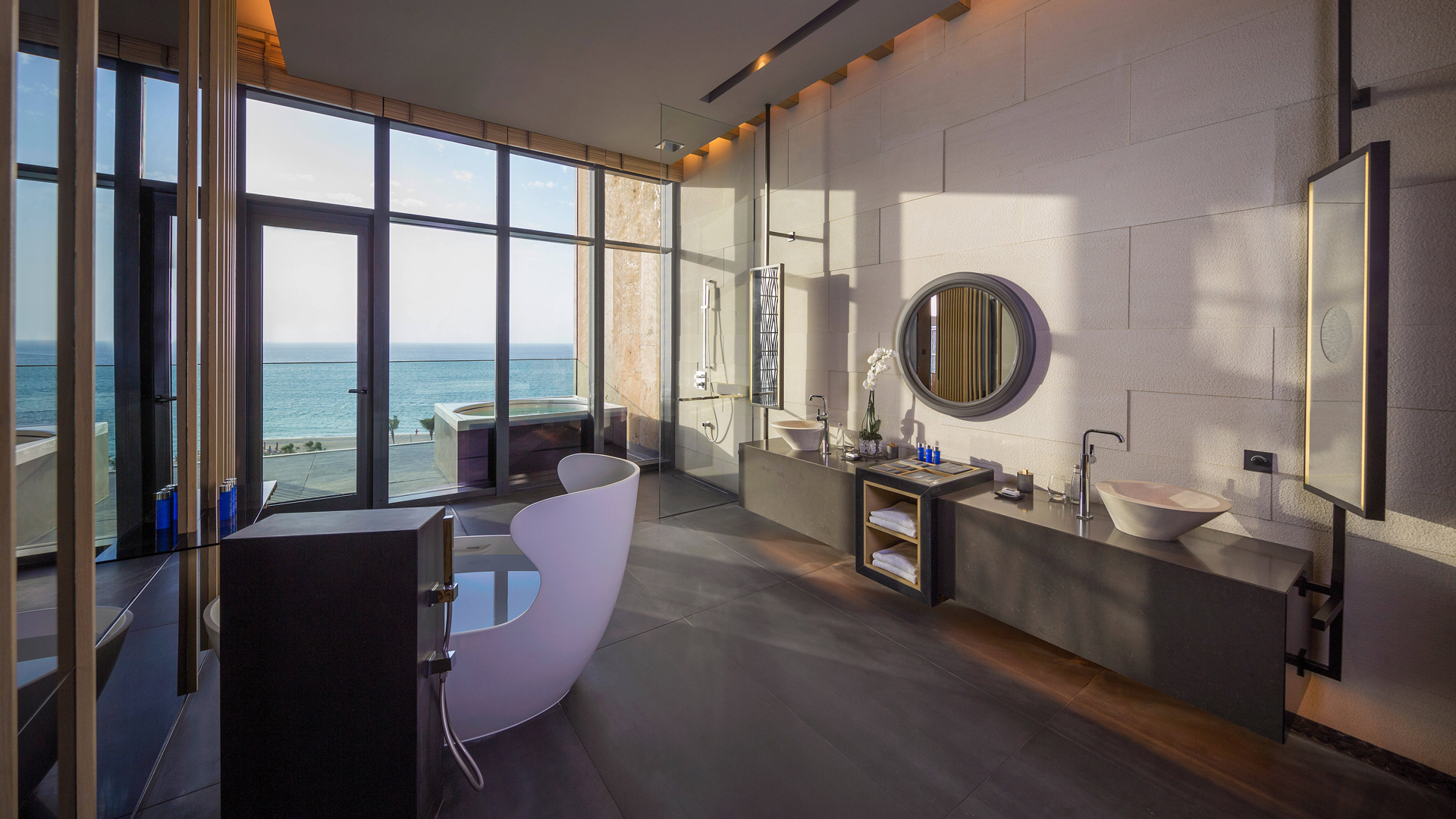 Spacious luxury bathroom with modern fixtures, including a freestanding white bathtub, dual vanities with vessel sinks, round mirrors, a glass-enclosed rain shower, and floor-to-ceiling windows offering breathtaking ocean views and access to an outdoor soaking tub.