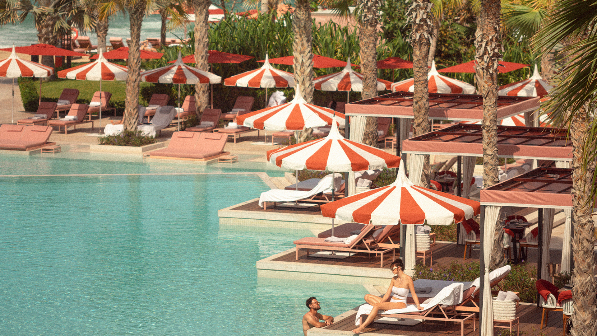 Summer Offer A luxurious poolside setting featuring striped red and white umbrellas, pink sun loungers, and cabanas surrounded by palm trees. A man is relaxing in the pool while a woman lounges on a chair under an umbrella, creating a vintage, summer vacation vibe.