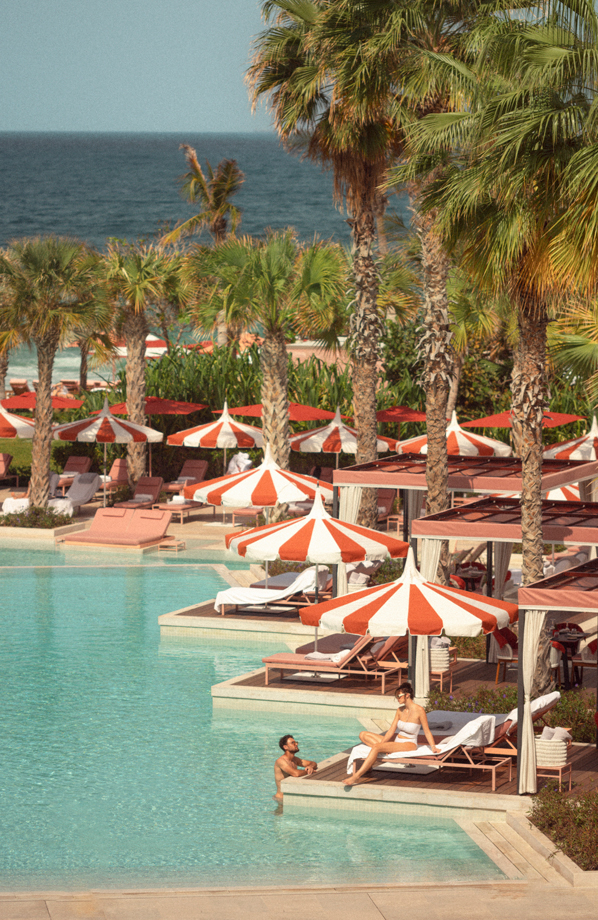 Luxurious beachfront poolside setting with red-and-white striped umbrellas, palm trees, and private cabanas. A man relaxes in the water while a woman lounges on a sunbed, both enjoying the sunny weather and ocean view in a stylish resort ambiance.