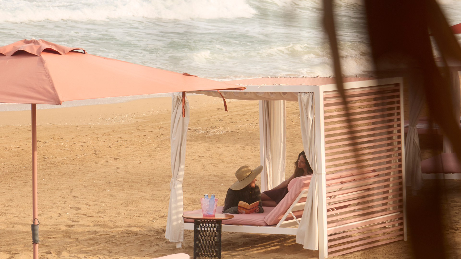 Tranquil beach scene with two women relaxing under a shaded cabana, surrounded by soft pink loungers and umbrellas. Gentle waves roll onto the sandy shore, with a distant view of waterfront buildings across the bay, framed by tropical foliage.