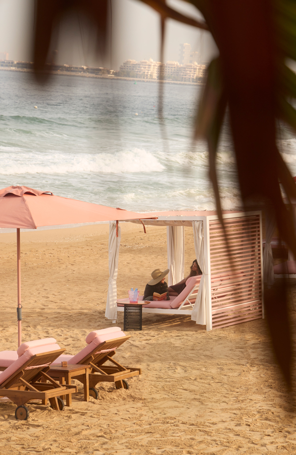 Tranquil beach scene with two women relaxing under a shaded cabana, surrounded by soft pink loungers and umbrellas. Gentle waves roll onto the sandy shore, with a distant view of waterfront buildings across the bay, framed by tropical foliage.