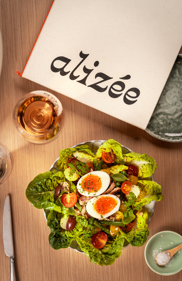 Top-down view of a vibrant salad with cherry tomatoes, crisp lettuce, green beans, herbs, and a soft-boiled egg, paired with a glass of rosé wine. A fabric napkin with 'Alizée' branding is partially visible on a wooden table, creating a fresh and inviting dining scene.