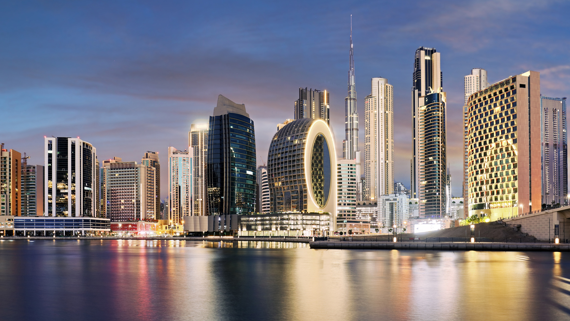 Stunning twilight view of Dubai's modern skyline reflected on the calm waters of Dubai Creek. Iconic buildings include the illuminated circular ME Dubai hotel and the towering Burj Khalifa, framed by a mix of contemporary high-rises under a colourful evening sky.