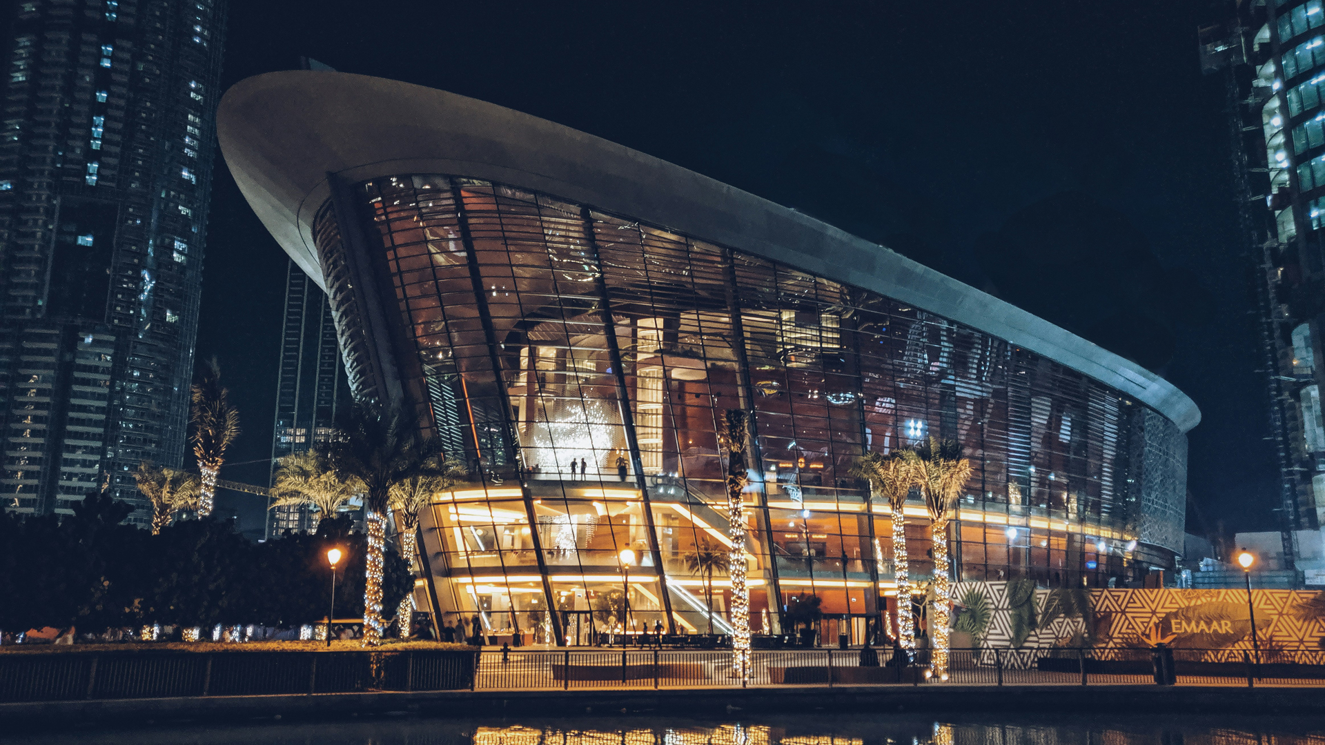 Night view of the Dubai Opera, a striking dhow-shaped architectural landmark with illuminated glass façade and modern interiors. The surrounding palm trees wrapped in lights and the city skyline in the background create an elegant cultural atmosphere.