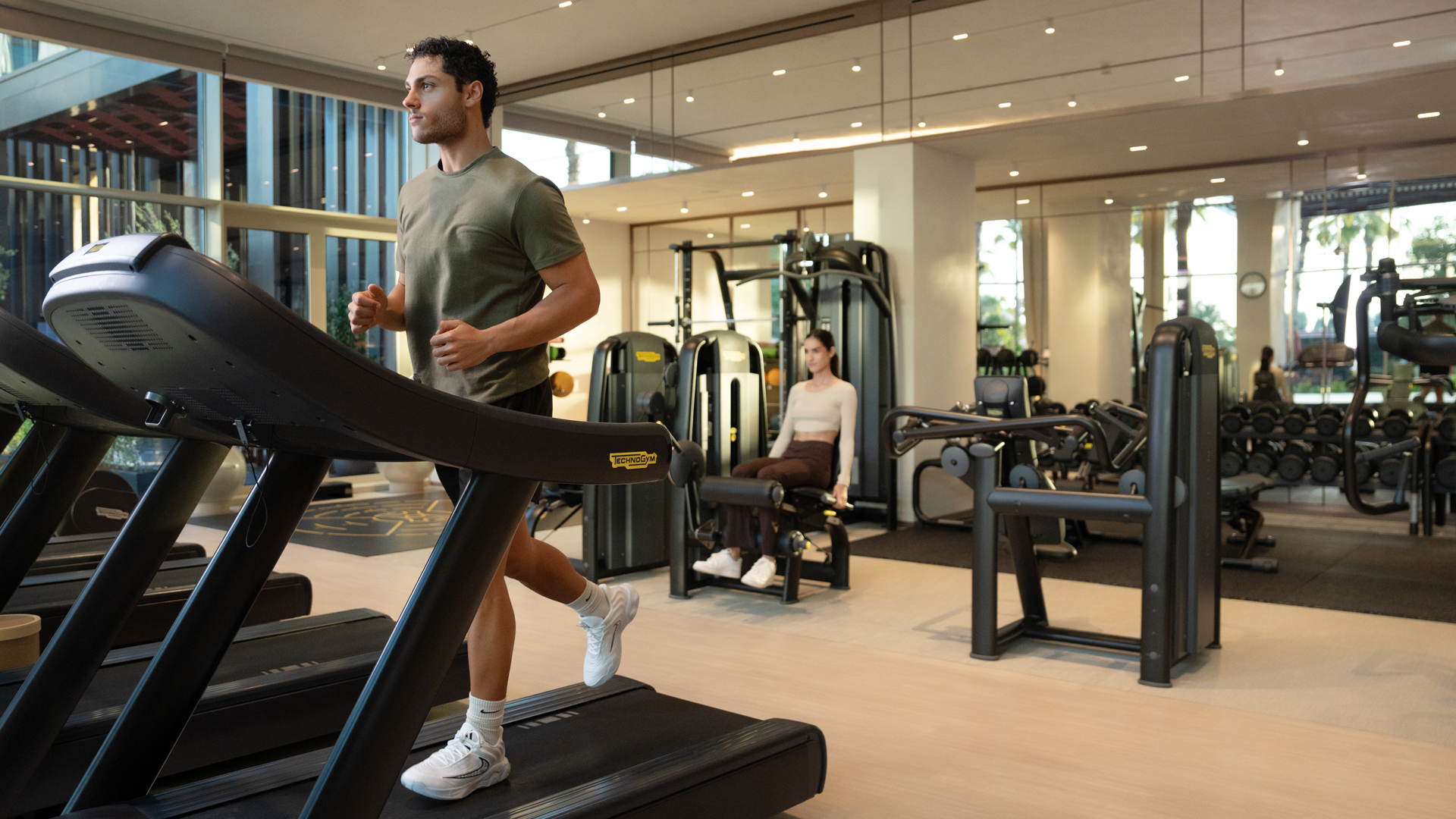 Guests working out at Banyan Tree Dubai gym—male running on treadmill and female using strength equipment, in a bright, fully equipped Technogym facility.